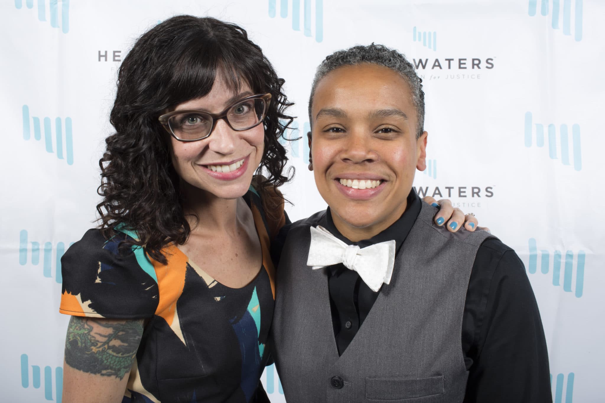 Two guests at the 2016 Change Makers Gala. One wears a multicolor, cap-sleeved dress and glasses; the other wears a black button up shirt, grey vest, and white bowtie. Both are smiling.